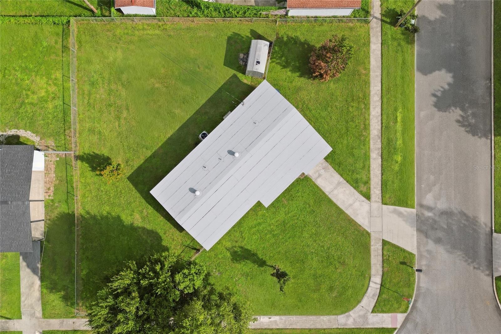 Aerial view of a home with a light roof, fenced lawn, and curved walkway beside the street.