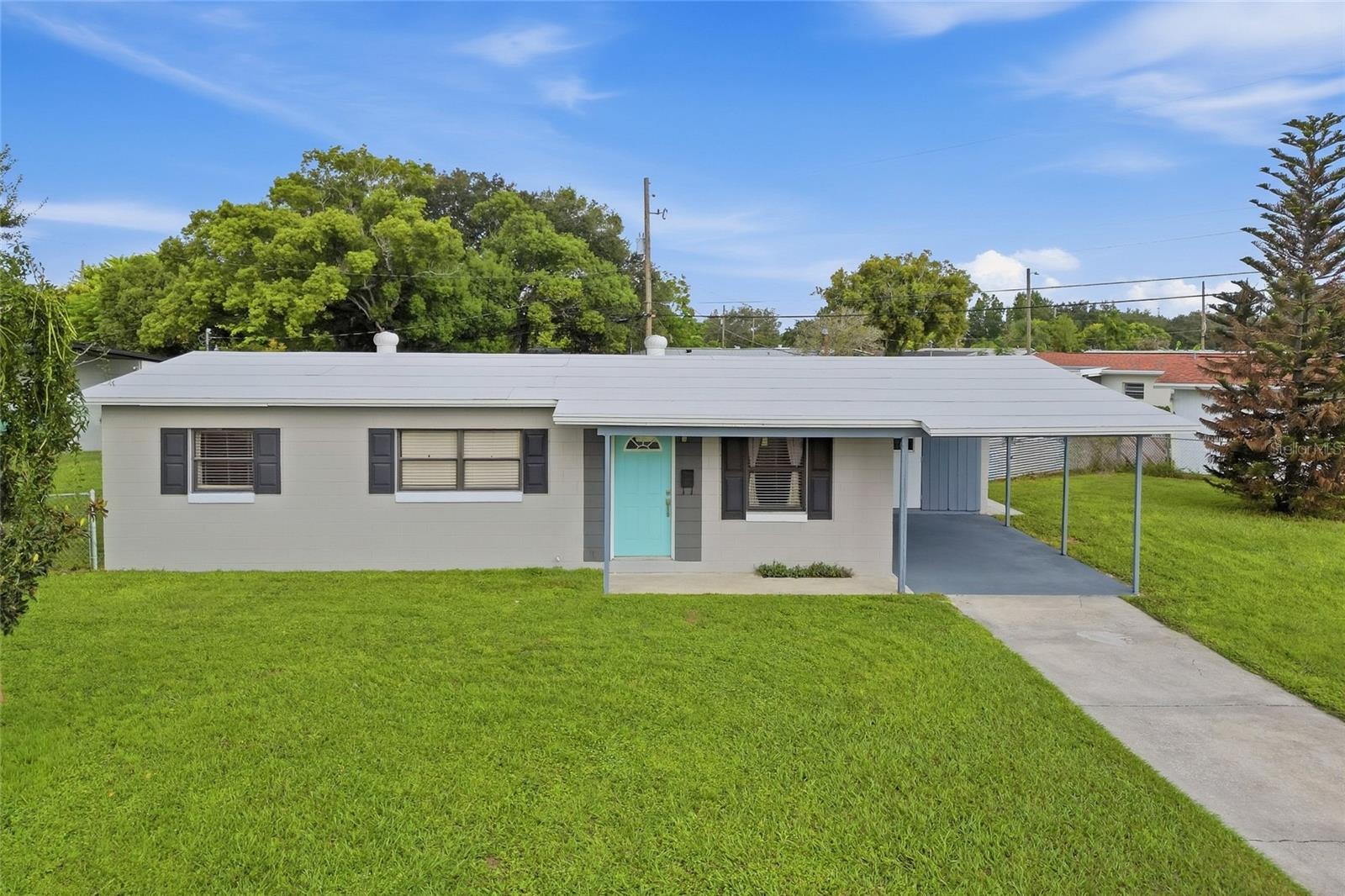 Single-story gray home with a turquoise front door, covered carport, and broad green lawn.