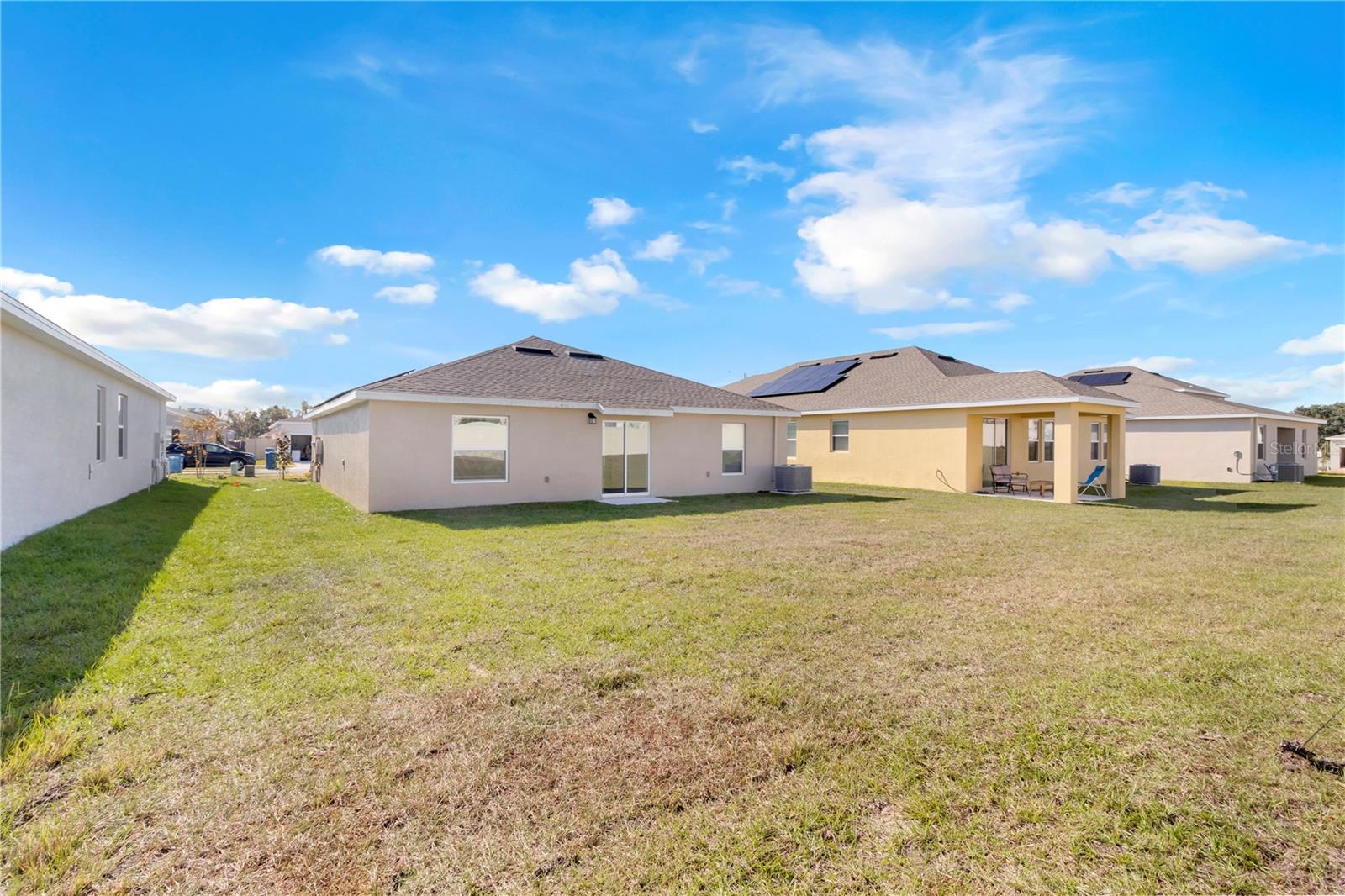 Rear of a single-story home overlooking a broad grassy backyard, with neighboring homes and solar panels nearby.
