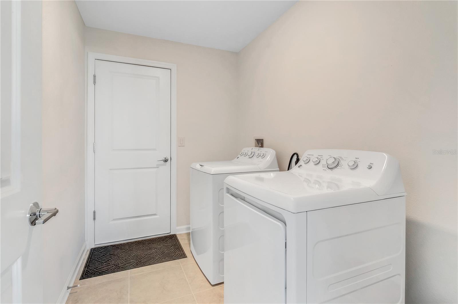 Laundry room with white washer and dryer, beige tile floor, and a dark mat by the door.