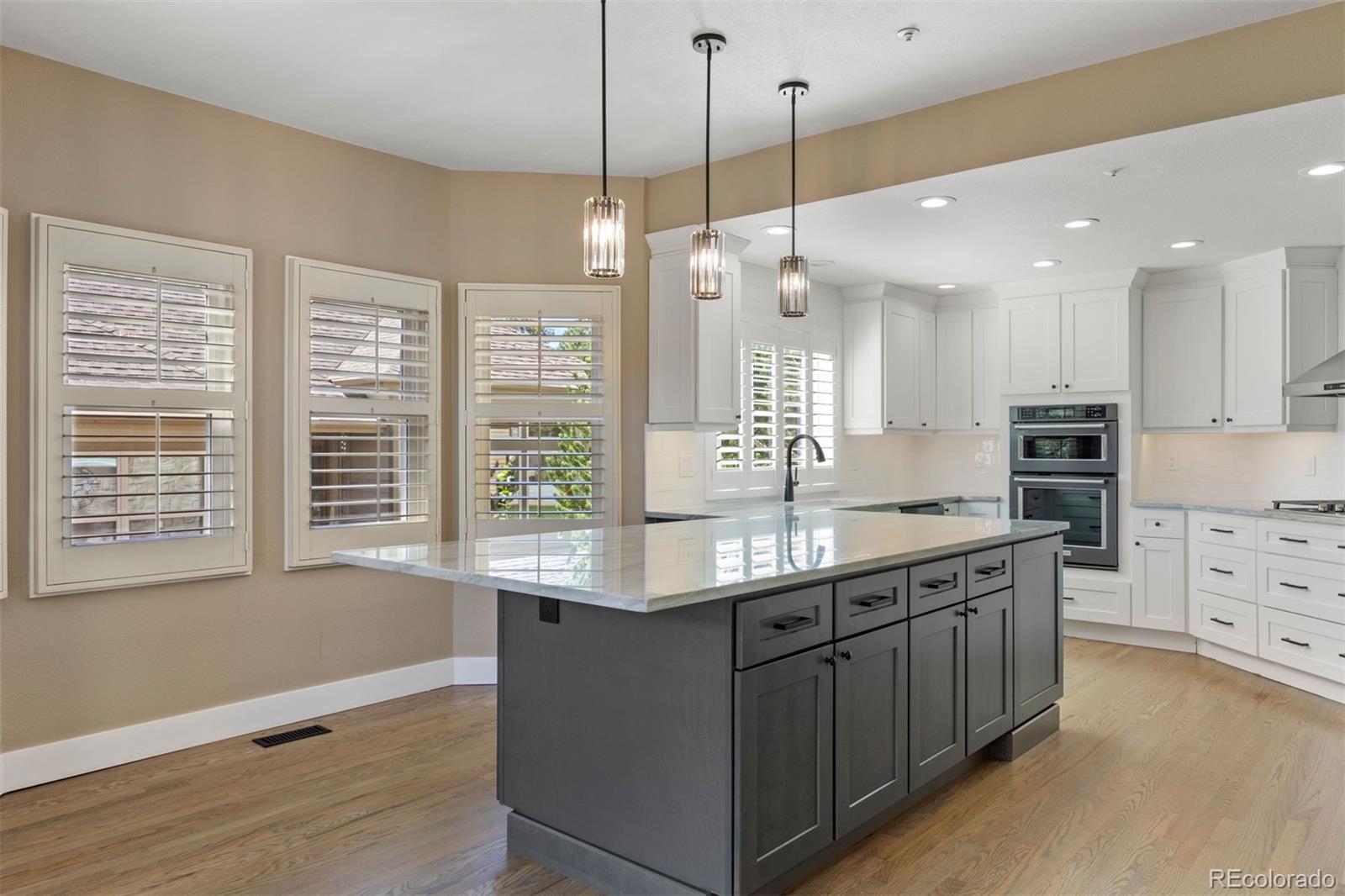 Kitchen with a large dark island, white cabinetry, double wall ovens, and plantation shutters.