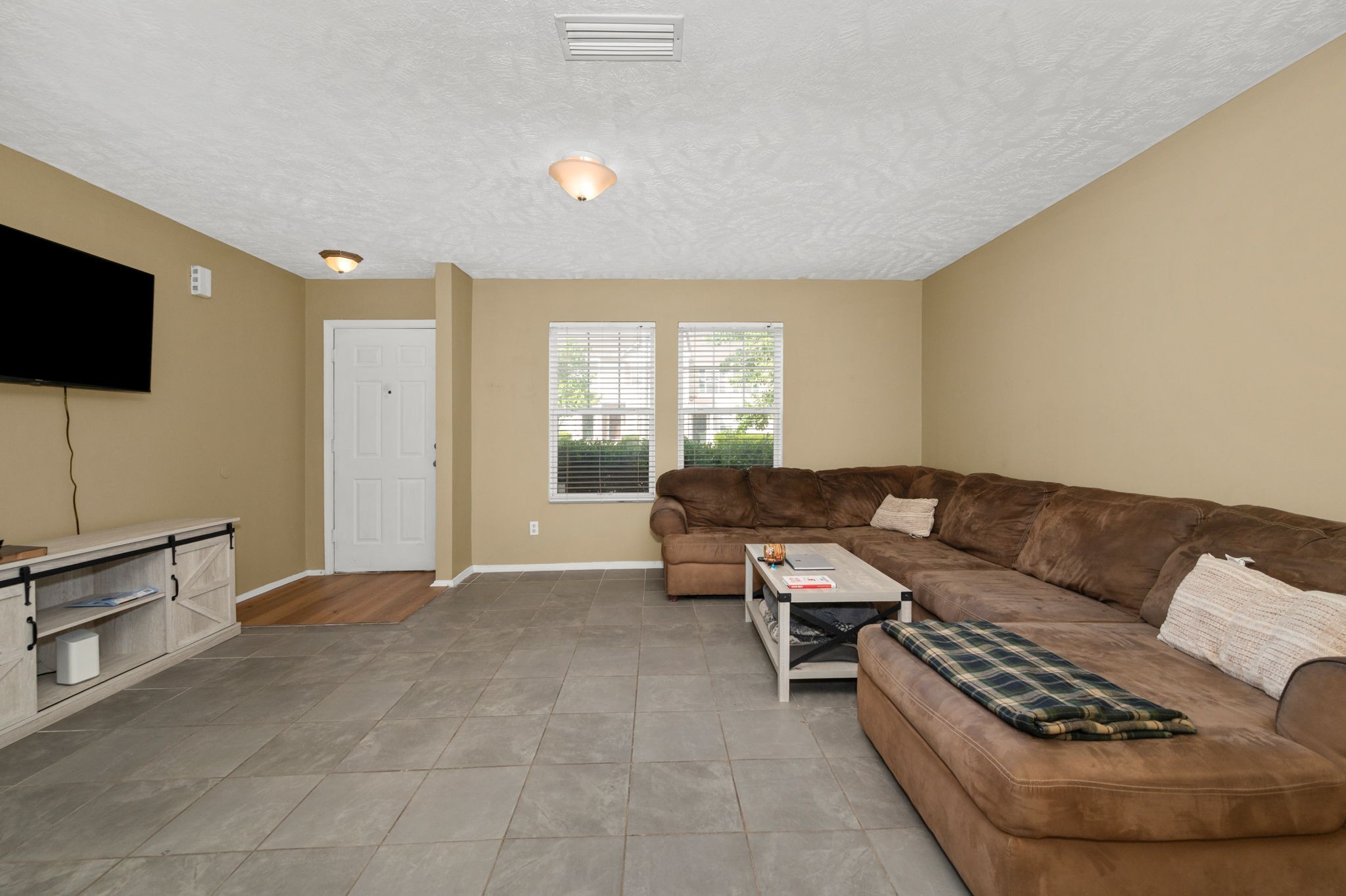 Living room with a brown sectional sofa, gray tile floors, and two windows with blinds.