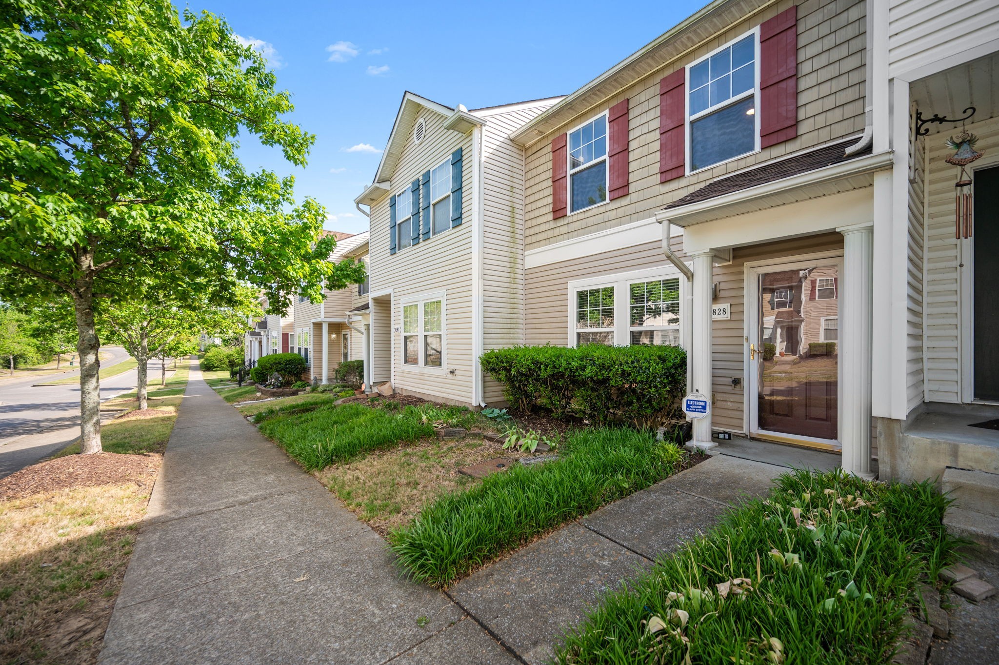Beige townhouse with red shutters, a covered entry, and landscaped walkway along a tree-lined sidewalk.
