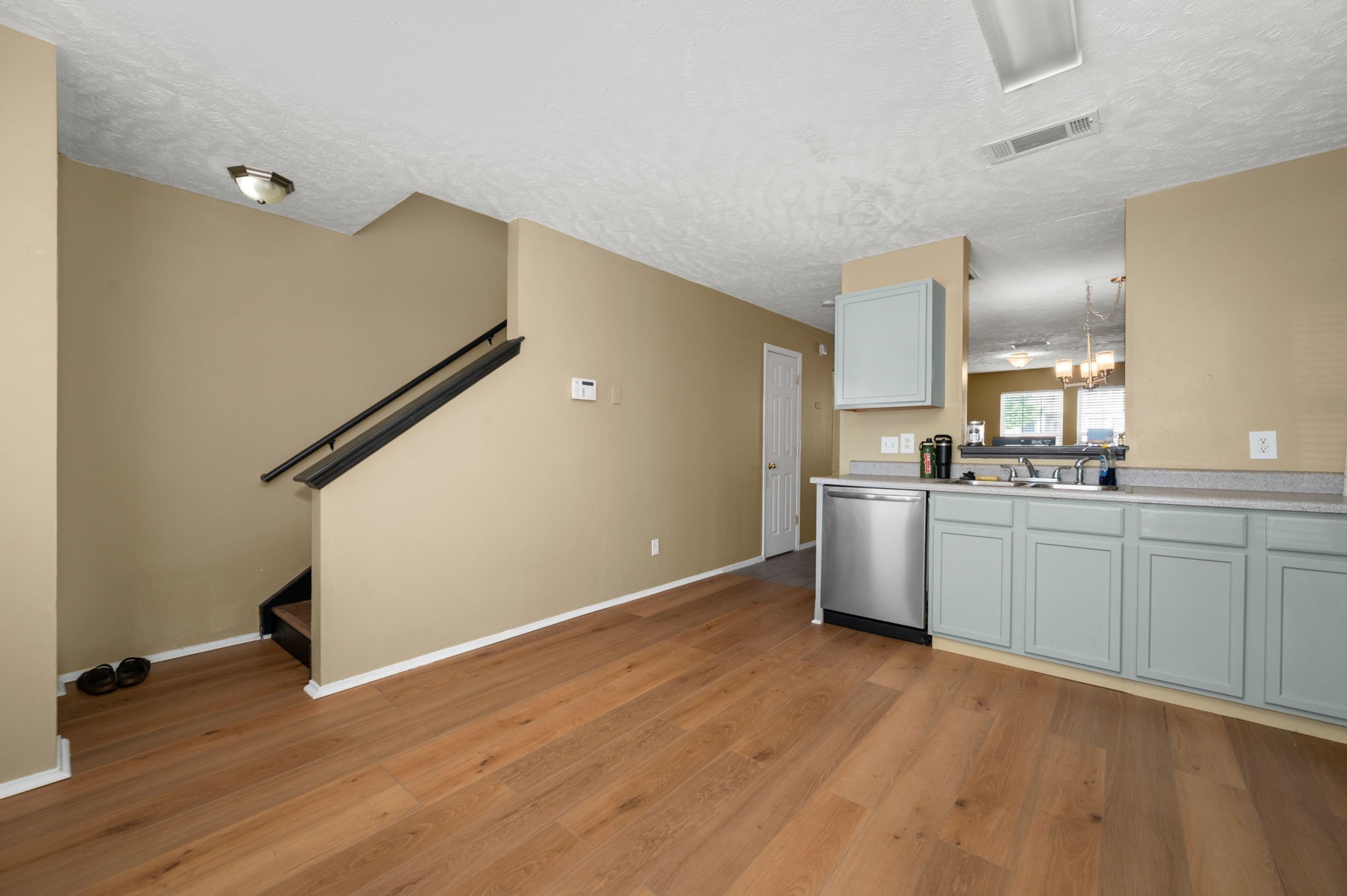 Kitchen with pale blue-gray cabinets, stainless dishwasher, wood flooring, and an adjacent staircase.