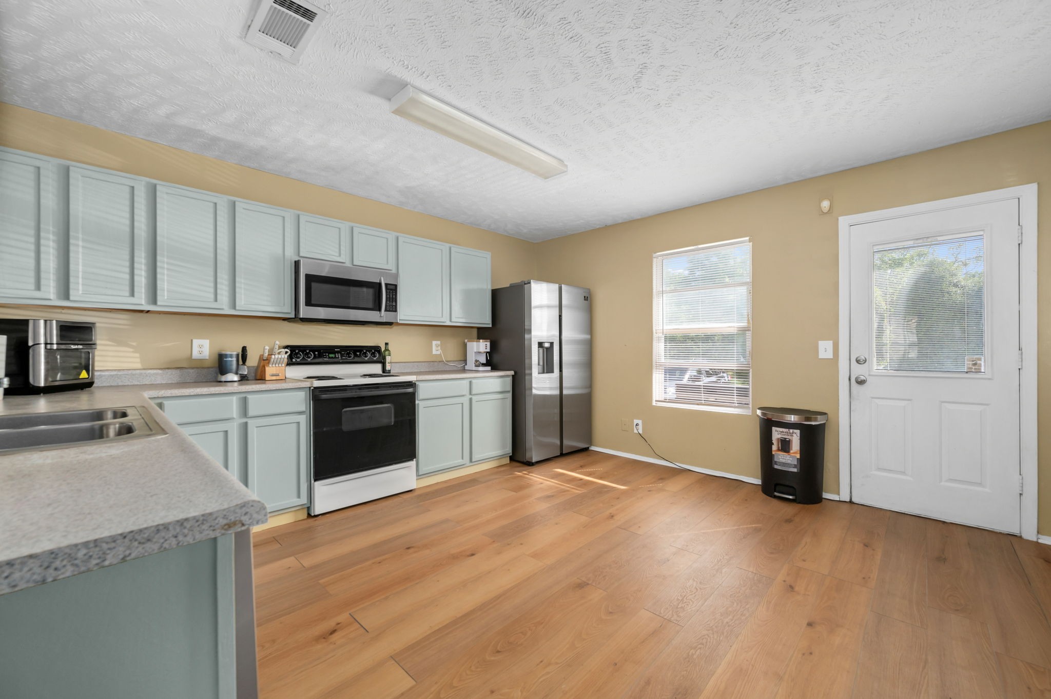 Kitchen with pale blue-green cabinets, stainless appliances, and wood flooring beside a window and exterior door.