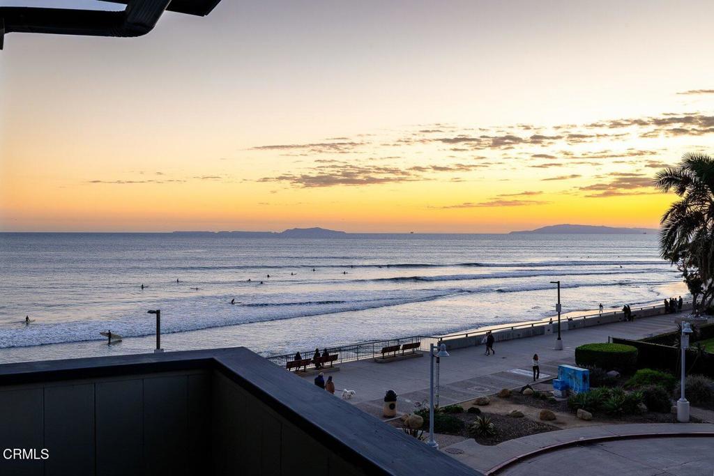Ocean view at sunset from a balcony, with surfers, a beachfront promenade, and distant islands.