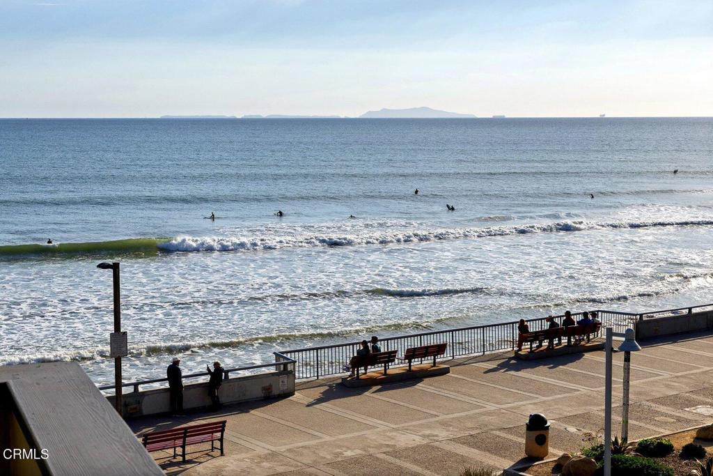 Ocean surf viewed from a seaside promenade with benches and surfers in the water.