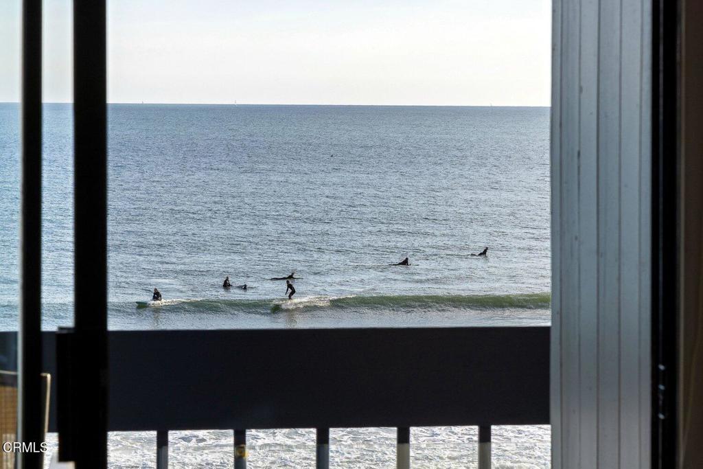 Ocean view with surfers riding small waves, framed by a doorway and balcony railing.