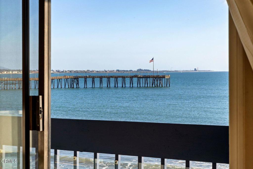 Ocean and long pier viewed from a balcony through an open sliding glass door.