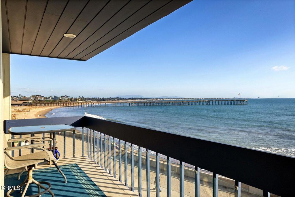 Oceanfront balcony with outdoor seating overlooking a sandy beach and long pier.