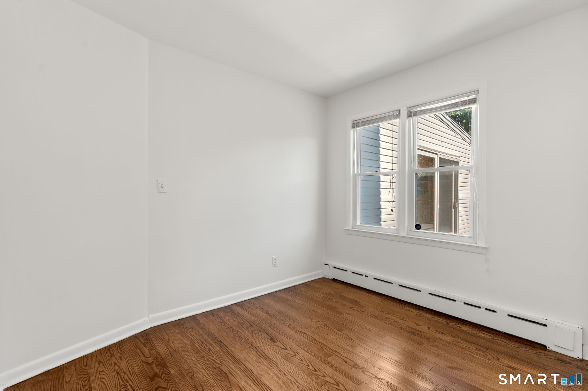 Vacant room with hardwood floors, white walls, and two windows above baseboard heating.