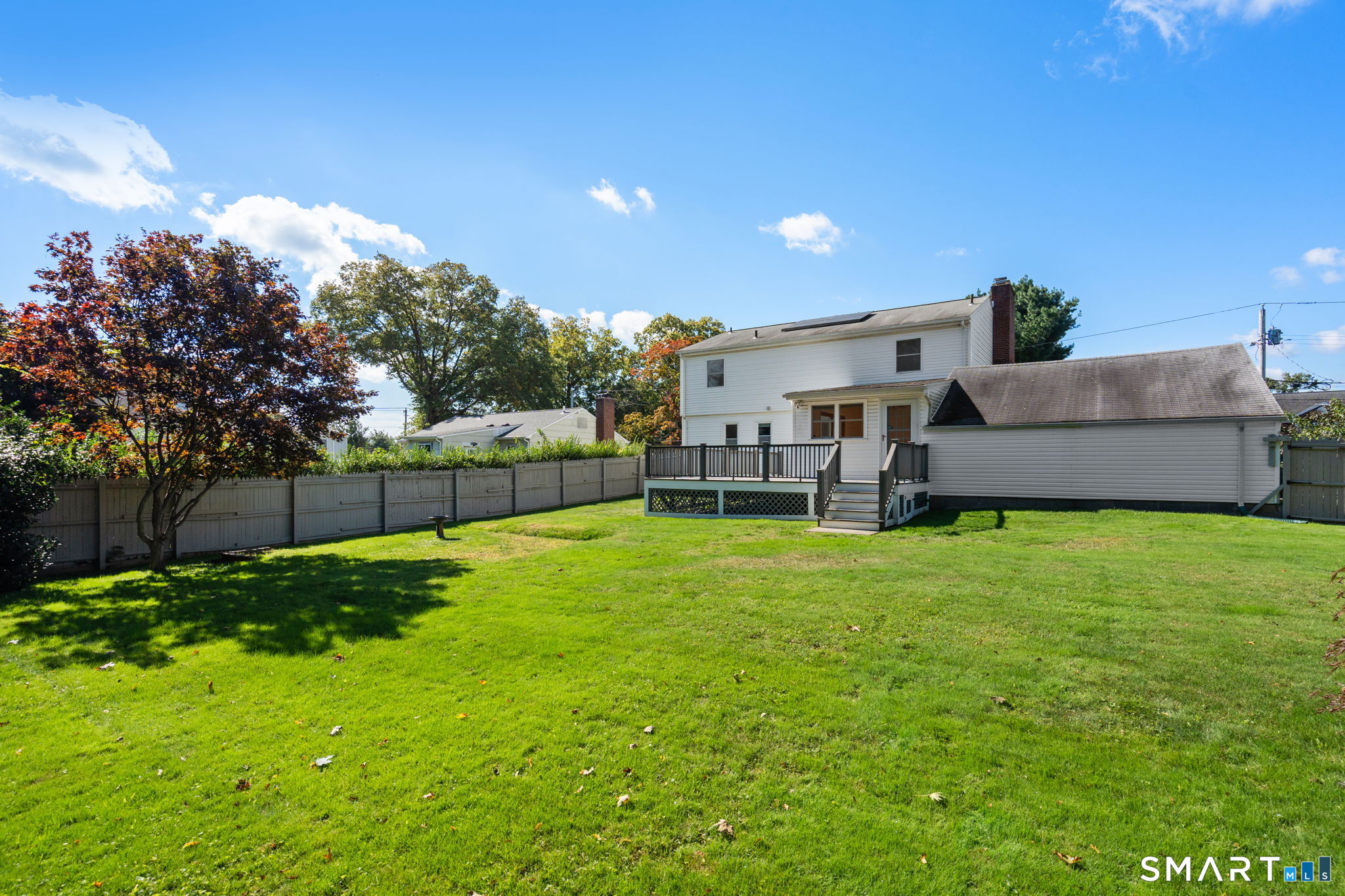 White two-story house with a raised rear deck, solar panels, and a large fenced lawn.