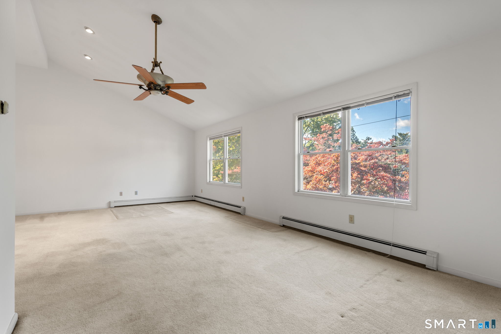 Vacant carpeted room with a ceiling fan and large windows overlooking autumn foliage.