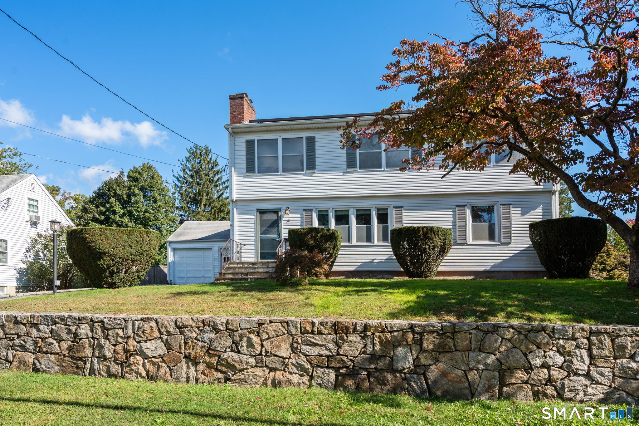 Two-story white-sided house with an attached garage, trimmed shrubs, and a stone retaining wall.