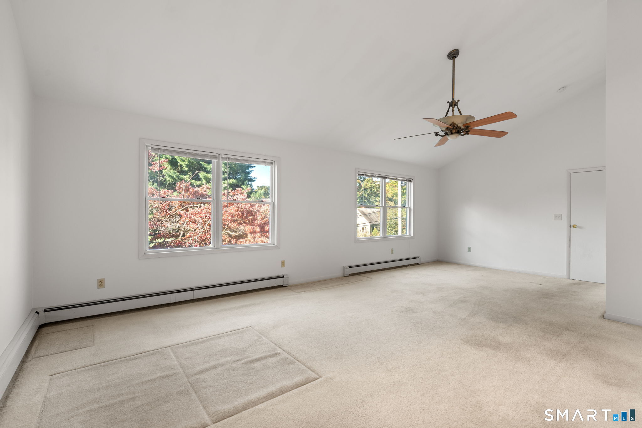 Empty carpeted room with two windows, baseboard heating, and a ceiling fan.