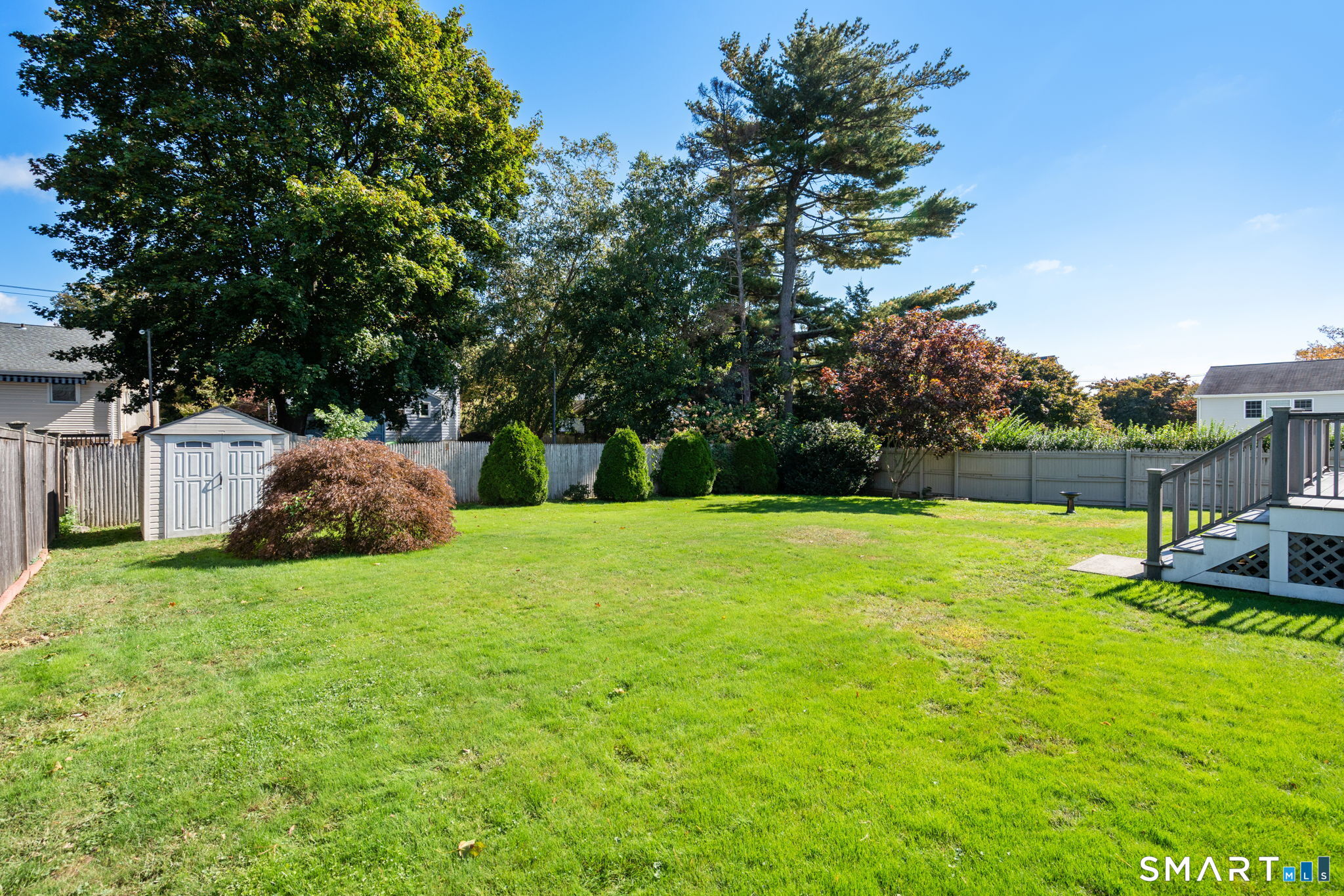 Spacious fenced backyard with a green lawn, mature trees, a shed, and deck stairs.