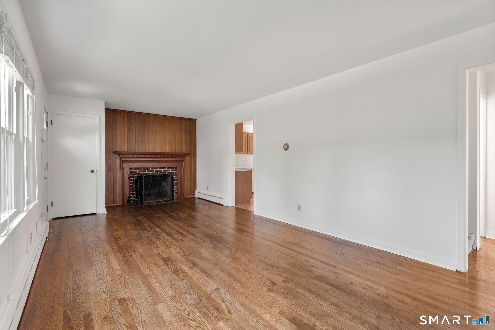 Empty living room with hardwood floors, large windows, and a wood-paneled fireplace.