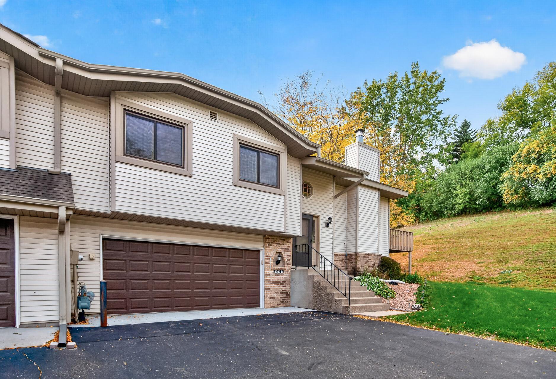 Two-story townhouse with a brown double garage door and front steps beside a sloping, tree-lined lawn.