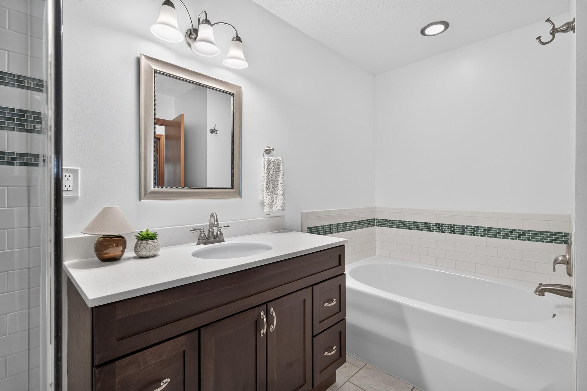 Bathroom with dark wood vanity, framed mirror, and bathtub bordered by white tile with a green accent band.