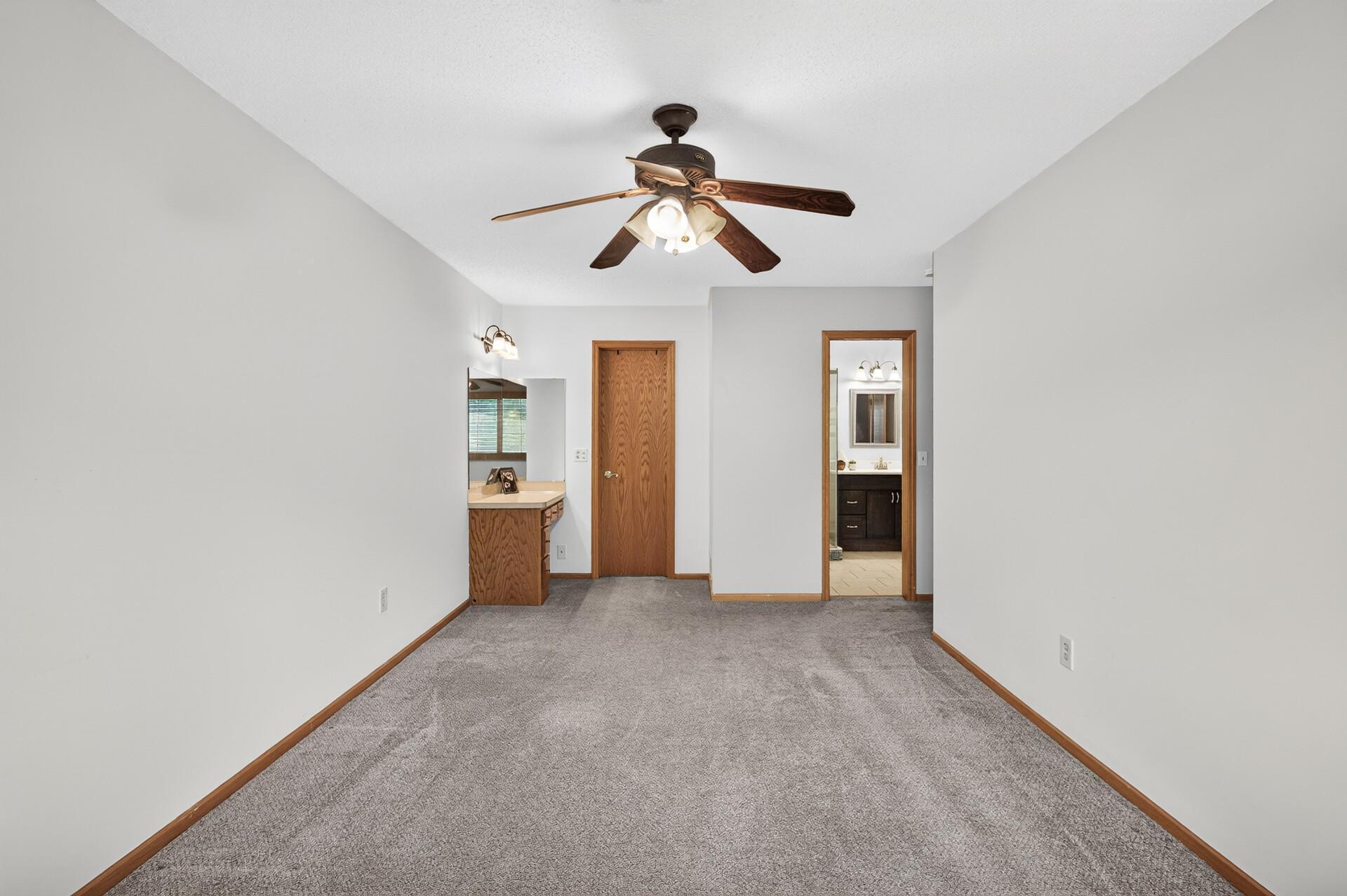 Empty carpeted bedroom with a wood vanity, ceiling fan, and bathroom doorway.