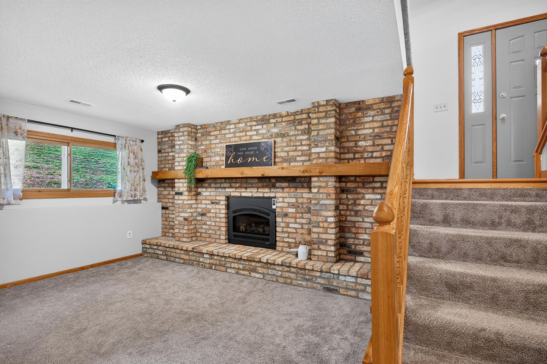 Living room with a brick fireplace, broad wood mantel, and carpeted staircase with wooden railing.