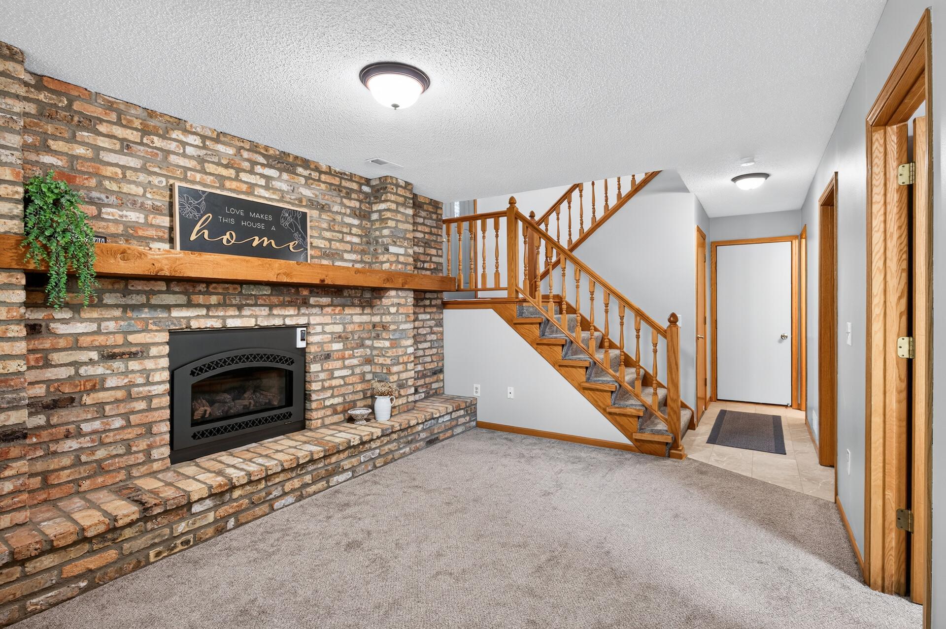 Carpeted living area with a brick fireplace and hearth beside a wooden staircase.