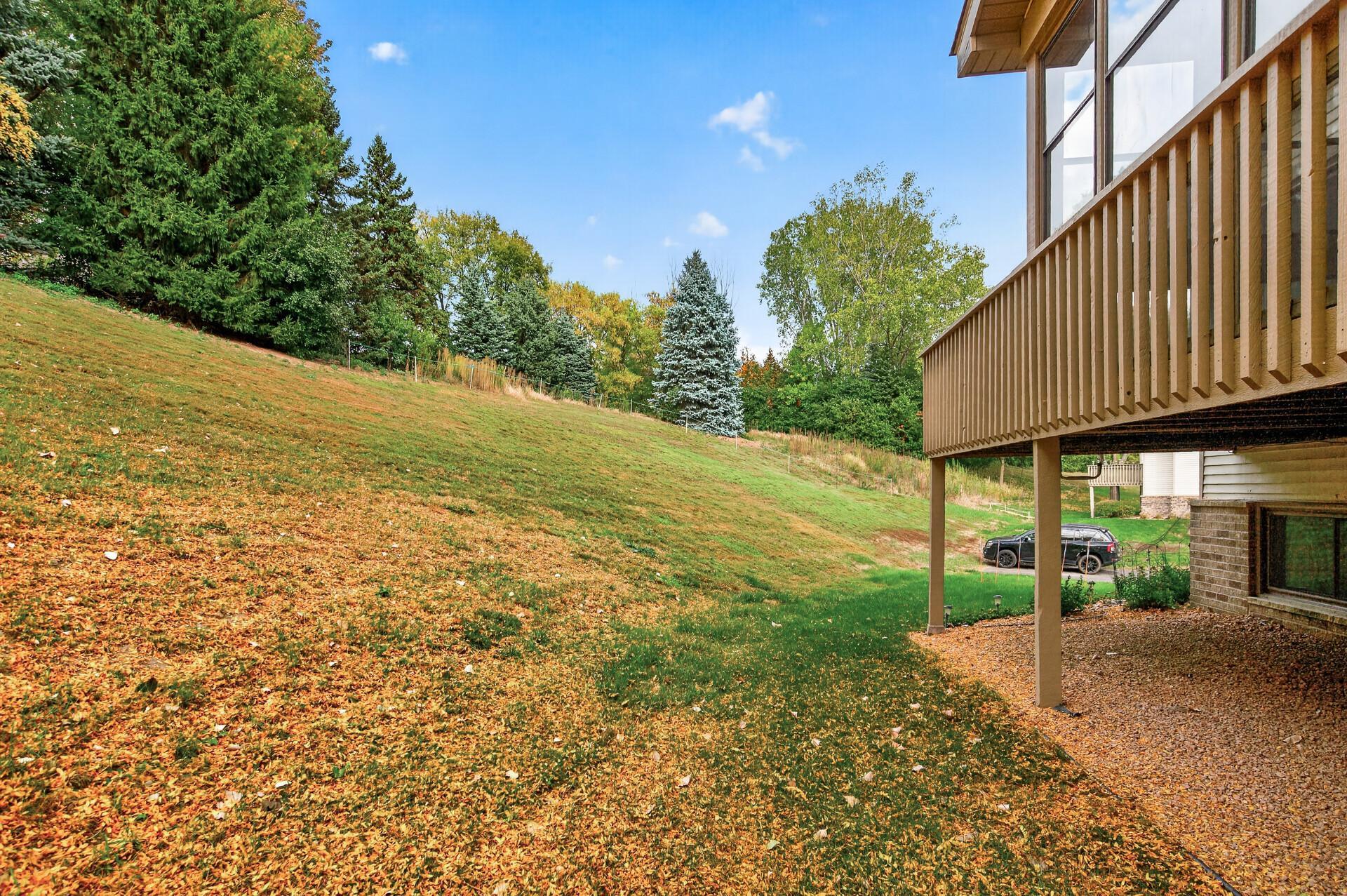 Raised wooden deck beside a sloping lawn covered with autumn leaves and mature trees.