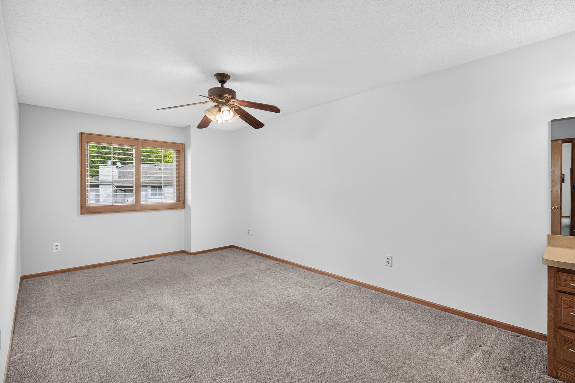 Empty carpeted bedroom with a ceiling fan, wood-trimmed window, and partial vanity at right.