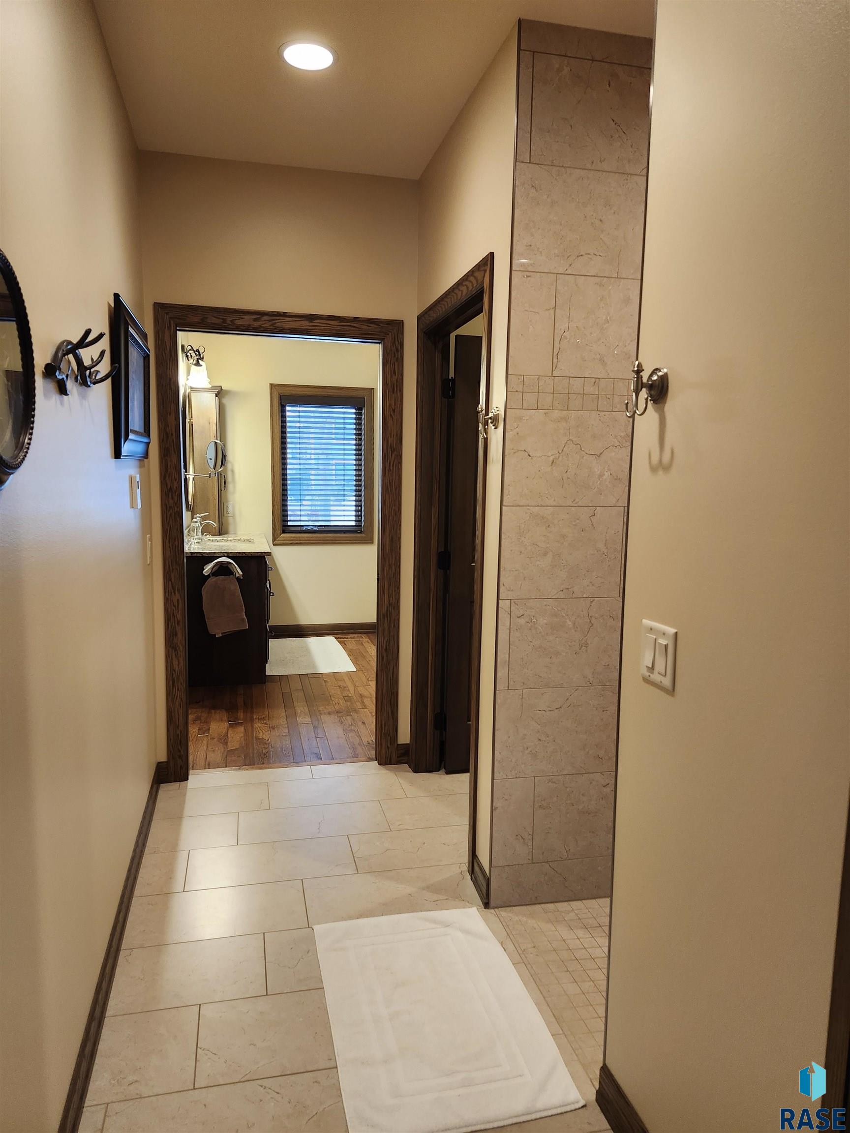 Bathroom with beige stone tile, a white bath mat, and a view through to the vanity and window.
