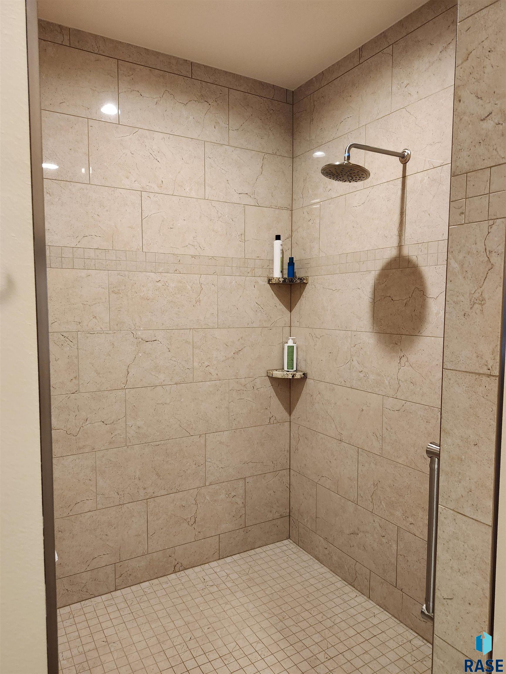 Walk-in shower with beige stone tile walls, rainfall showerhead, and corner shelves.