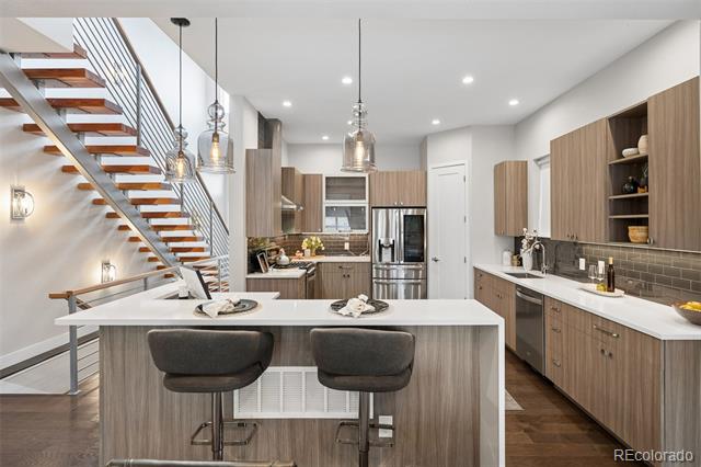 Modern kitchen with a large island, wood cabinetry, stainless steel appliances, and an open-riser staircase.