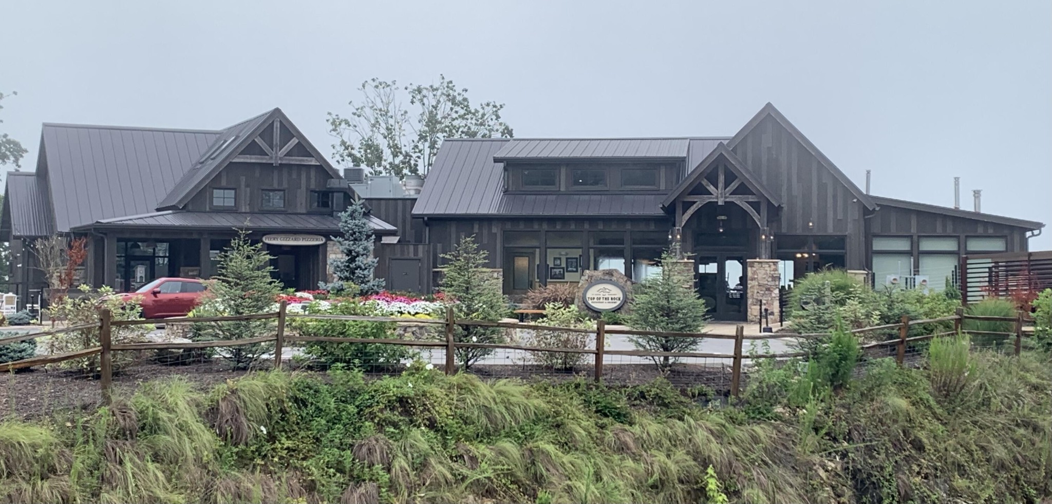 Dark timber building with metal gable roofs, stone entrance, and landscaped grounds behind a split-rail fence.