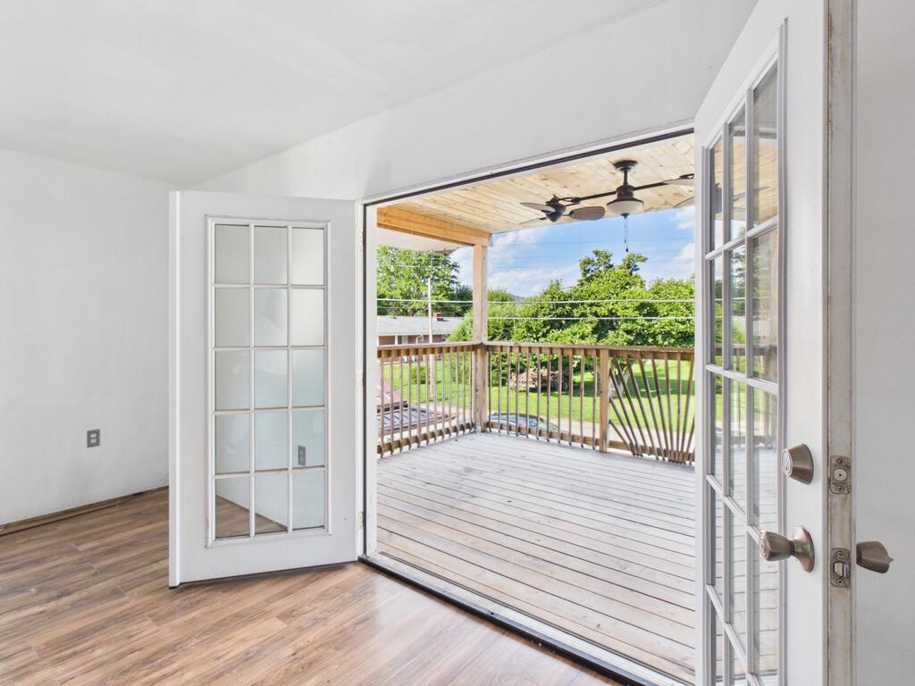 Empty room opens through French doors onto a covered wooden deck overlooking a green yard.