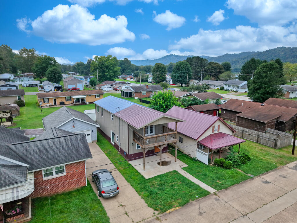 Aerial view of a two-story home with a covered balcony and carport, surrounded by neighboring homes and wooded hills.