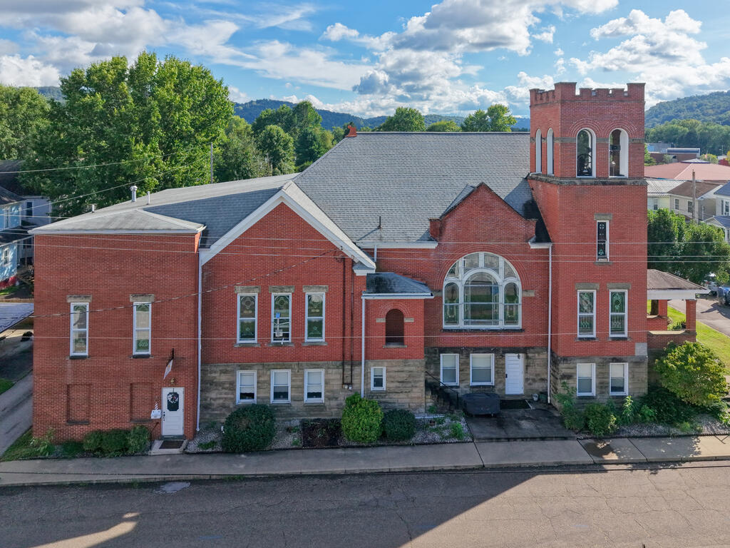 Red-brick building with a square tower, arched windows, and wooded hills behind it.