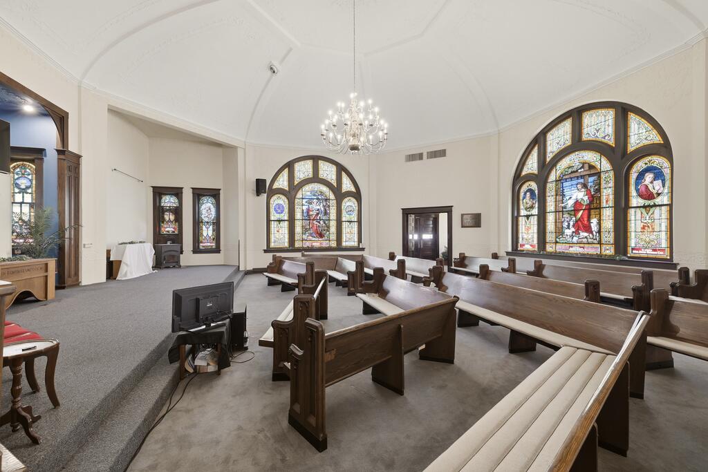 Chapel-like room with wooden pews, arched stained-glass windows, and a raised platform.