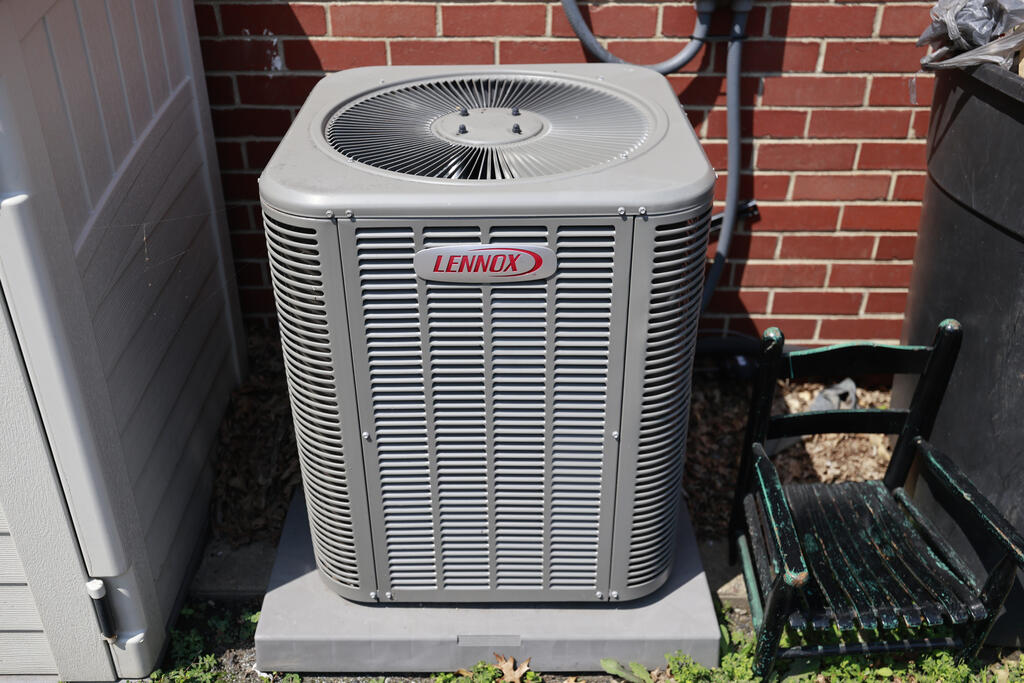 Outdoor Lennox air-conditioning unit beside a brick wall and weathered chair.