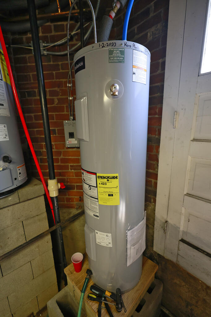 Electric water heater in a basement utility area, backed by exposed brick and plumbing.