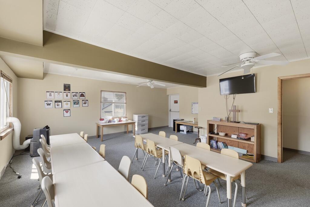Classroom-like room with rows of tables and chairs, framed pictures, and a wall-mounted television.