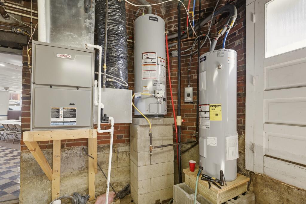 Utility area with a Lennox furnace, two water heaters, and exposed pipes against brick walls.