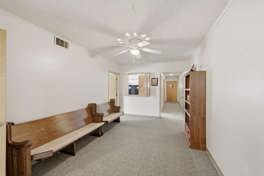 Interior hallway with wooden waiting benches, carpet, a ceiling fan, and a service window.