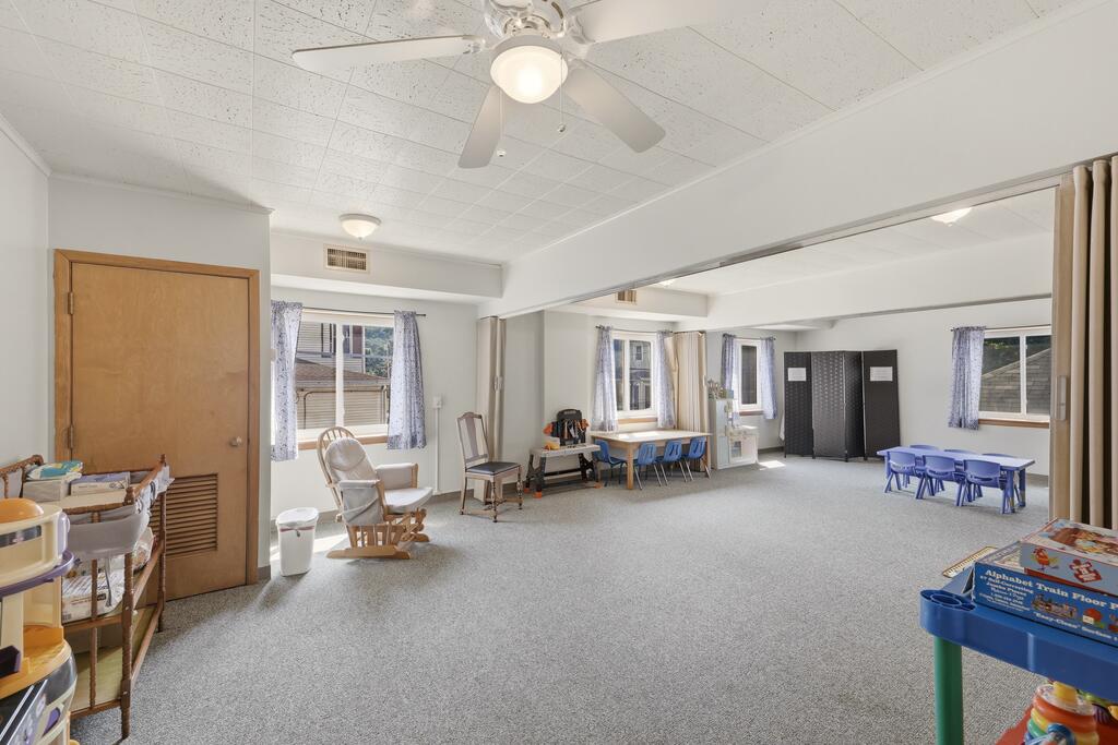 Carpeted classroom with a ceiling fan, windows, small tables and chairs, and children’s play furniture.