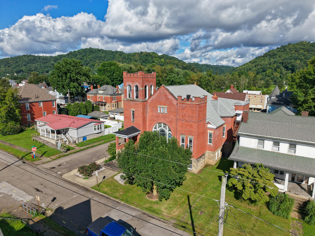 Red-brick building with a square tower among homes, with wooded hills in the background.