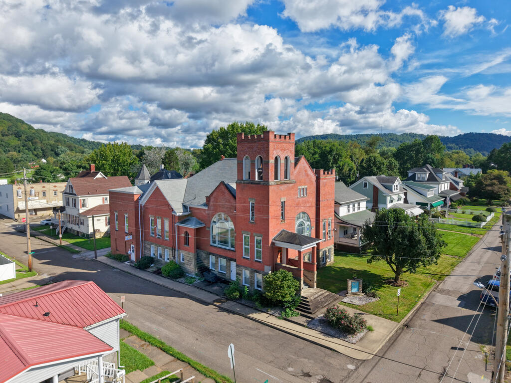 Red-brick building with a tall central tower at a street corner, surrounded by homes and wooded hills.