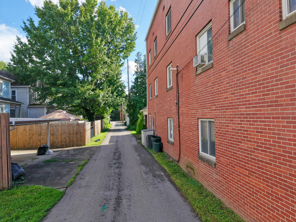 Red brick building beside a narrow paved alley, with a wooden fence and large leafy tree.