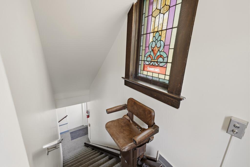Carpeted staircase with a brown stair lift beneath a colorful stained-glass window.