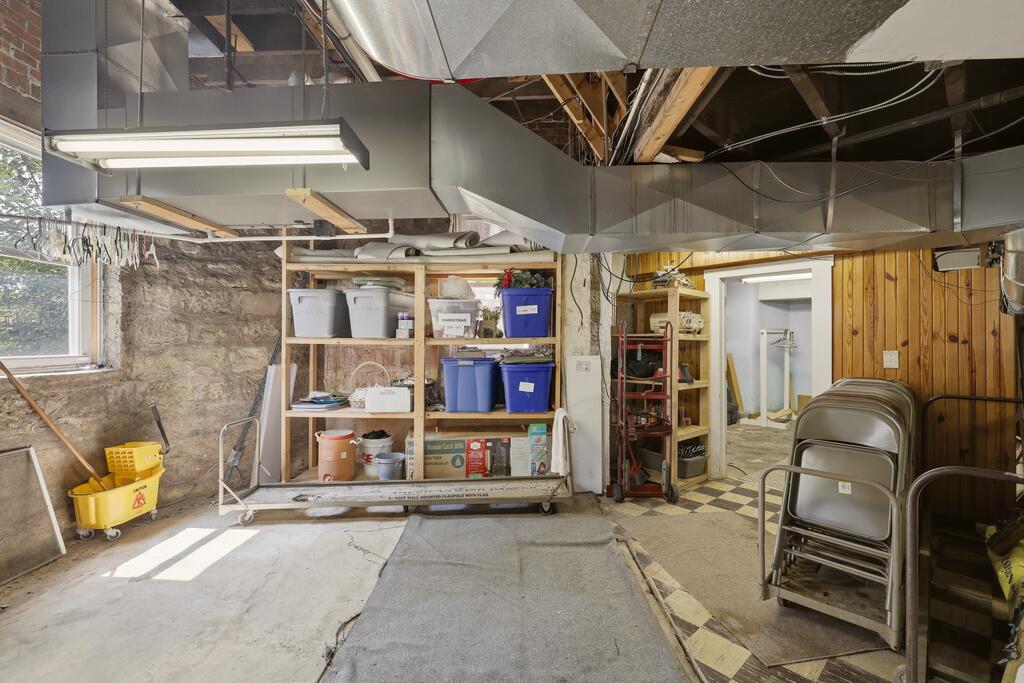Unfinished basement storage area with exposed ductwork, stone walls, shelving, and folding chairs.