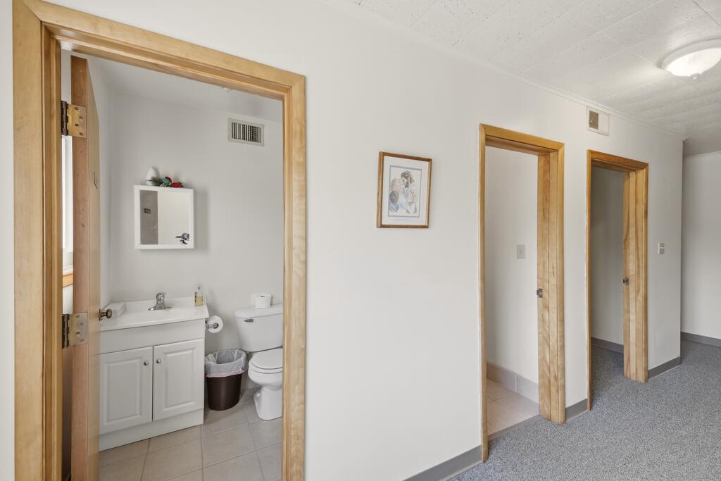 Hallway with light wood trim and gray carpet, looking into a bathroom with a white vanity and toilet.