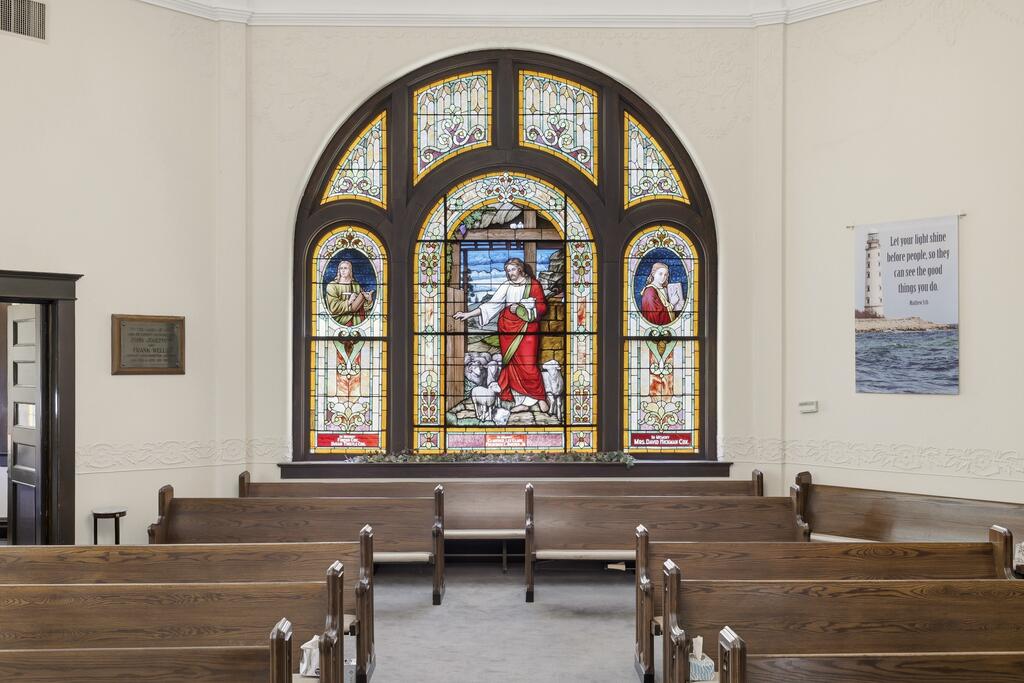 Chapel interior with wooden pews facing a large, arched stained-glass window.