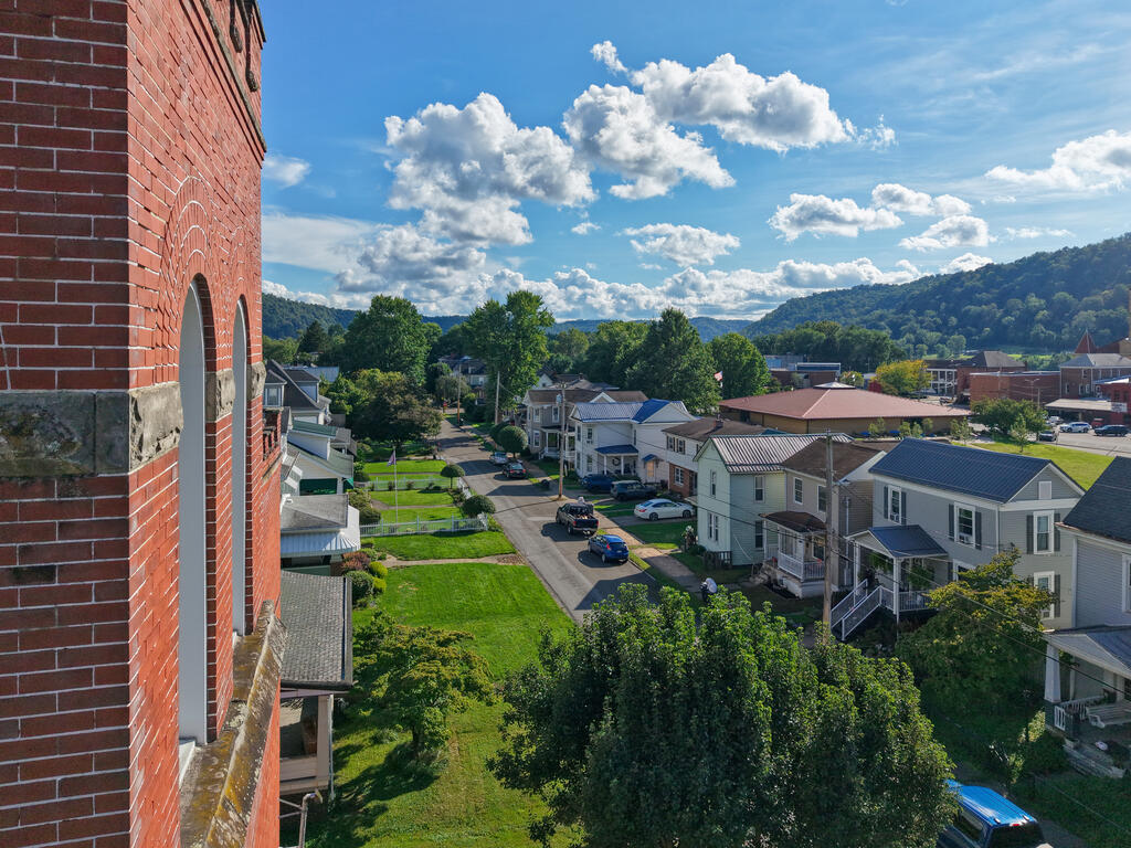 Residential street and houses viewed beside a red brick building, with wooded hills in the distance.